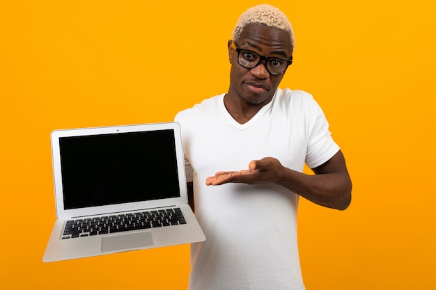 Afrikanischer Geschäftsmannmann in den Gläsern und im weißen T-Shirt hält einen Laptop mit einem Modell und einem gelben Studiohintergrund