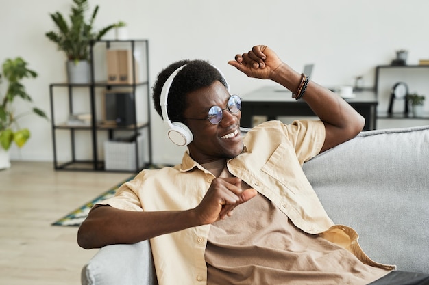 Afrikanischer glücklicher Mann in drahtlosen Kopfhörern, der auf dem Sofa tanzt und die Musik zu Hause genießt