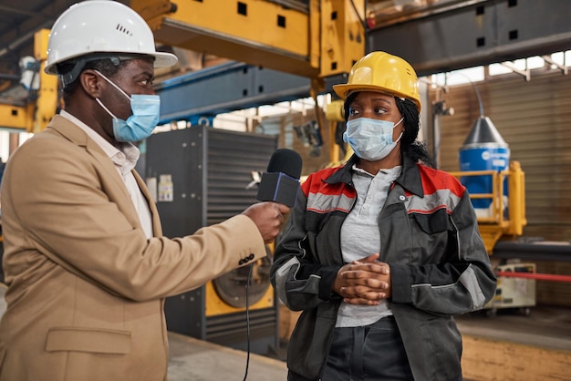 Afrikanischer Journalist in Maske interviewt den Arbeiter in Uniform in der Fabrik während einer Pandemie