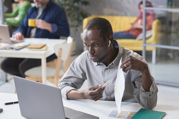 Afrikanischer junger Geschäftsmann, der auf ein Dokument in seinen Händen zeigt, während er eine Online-Konferenz auf dem Laptop hat