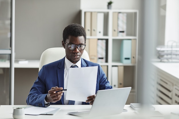 Afrikanischer junger Geschäftsmann in Brillen, die an seinem Arbeitsplatz vor Laptop sitzen und einen Vertrag lesen