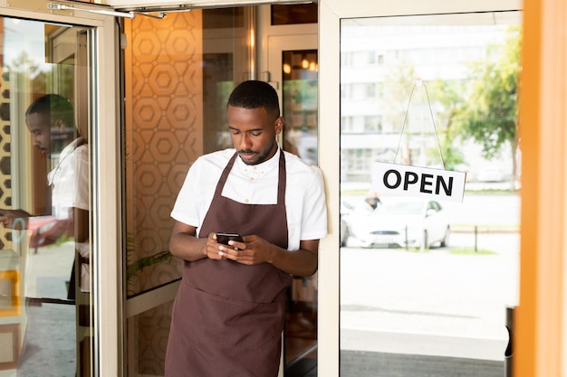 Afrikanischer junger Kellner in Uniform, der in der Tür eines Cafés oder Restaurants steht und neue Bestellungen von Kunden im Smartphone durchblättert