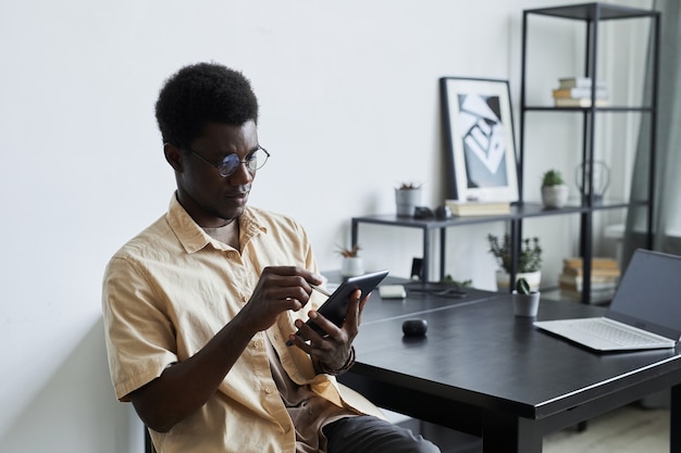 Afrikanischer junger Mann, der an seinem Arbeitsplatz sitzt und auf einem digitalen Tablet tippt, das er online arbeitet