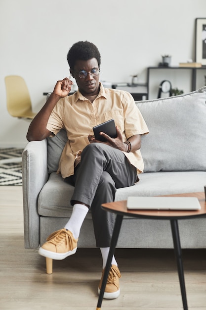 Afrikanischer junger Mann, der auf dem Sofa sitzt und während seiner Freizeit zu Hause ein digitales Tablet benutzt