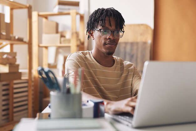 Afrikanischer junger Mann in Brillen, die am Tisch sitzen und online am Laptop im Büro arbeiten