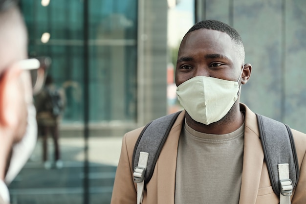 Afrikanischer junger Mann in der Schutzmaske, der mit seinem Kollegen spricht, während sie in der Stadt stehen