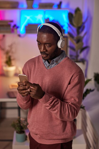 Afrikanischer junger Mann in drahtlosen Kopfhörern, die Musik online auf seinem Handy hören