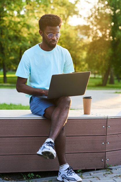 Afrikanischer junger Mann in Freizeitkleidung, der mit Laptop auf den Knien sitzt und im Freien online arbeitet