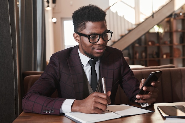 Afrikanischer junger Mann mit Brille, der online auf seinem Handy arbeitet und sich Notizen im Notizbuch macht, das im Restaurant sitzt