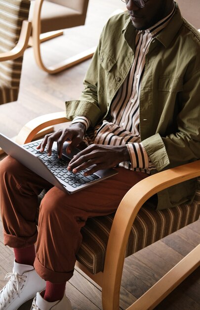 Afrikanischer junger Mann sitzt auf einem Stuhl und tippt auf einem Laptop, er arbeitet online im Büro