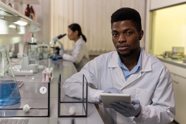 Afrikanischer junger Wissenschaftler, der bei der Arbeit an einem digitalen Tablet in die Kamera schaut und während des wissenschaftlichen Experiments zwei Arten von Mäusen untersucht