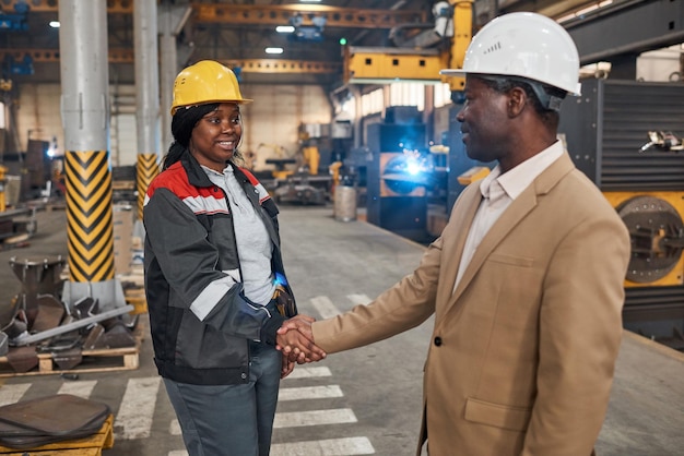 Afrikanischer Manager im Anzug schüttelt Arbeiterin in Uniform die Hand, um sie in der Fabrik einzustellen