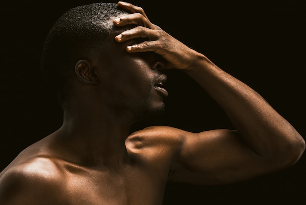 Afrikanischer Mann bedeckte sein Gesicht mit seiner Hand, Profilansicht eines nackten afroamerikanischen jungen Mannes, der auf schwarzem Hintergrund aufwirft