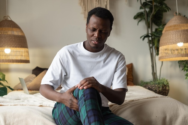 Afrikanischer Mann, der unter Knieschmerzen leidet, sitzt auf einem Sofa