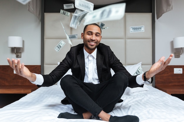 Afrikanischer Mann im Anzug wirft das Geld und sitzt auf dem Bett im Hotelzimmer