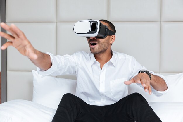 Afrikanischer Mann im Hemd unter Verwendung des virtuellen Realitätsgeräts, das auf Bett im Hotel sitzt