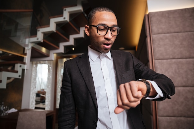 Afrikanischer Mann in Brille und Anzug mit Blick auf die Armbanduhr im Hotel