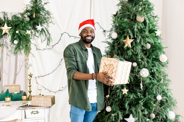 Afrikanischer Mann in der Weihnachtsmütze mit Geschenkbox auf dem Hintergrund des Weihnachtsbaumes