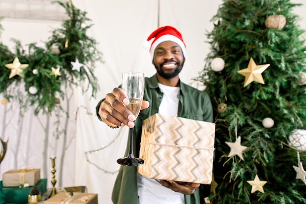 Afrikanischer Mann in der Weihnachtsmütze nahe Weihnachtsbaum, der Champagner und Geschenk verschenkt