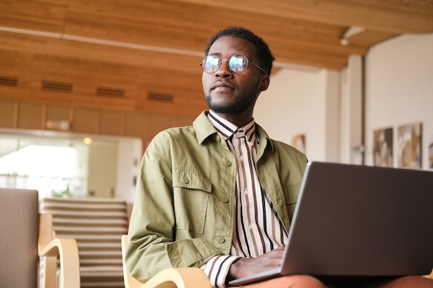 Afrikanischer Mann mit Brille, der online am Laptop arbeitet, während er im Café sitzt