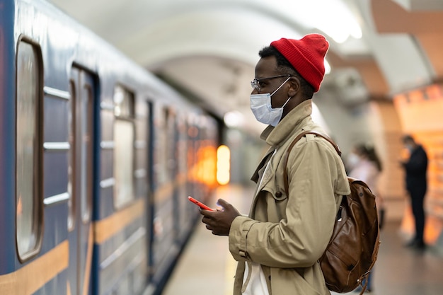 Afrikanischer Mann mit Gesichtsmaske in der U-Bahnstation wartet auf den Zug, indem er die sozialen Medien mit dem Smartphone scrollt