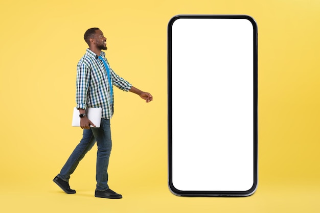 Afrikanischer Mann mit Laptop zu Fuß in der Nähe von großen Handy gelben Hintergrund