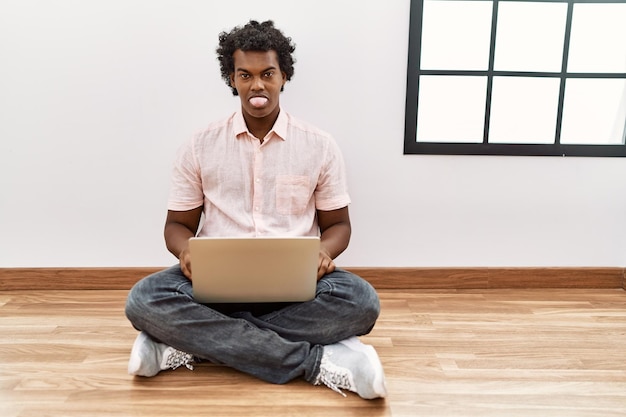 Afrikanischer Mann mit lockigem Haar, der einen Laptop benutzt, der auf dem Boden sitzt und die Zunge herausstreckt, glücklich über das Konzept des lustigen Gesichtsausdrucks