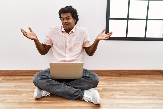 Foto afrikanischer mann mit lockigem haar, der einen laptop benutzt und ahnungslos auf dem boden sitzt, und sein verwirrter gesichtsausdruck mit armen und händen lässt zweifel am konzept aufkommen