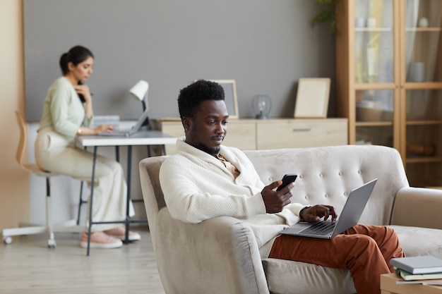 Afrikanischer Mann sitzt auf Sofa mit Laptop und verwendet Handy mit Frau, die in der Wand zu Hause arbeitet