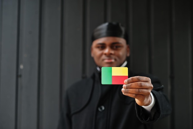 Afrikanischer mann trägt schwarzen durag, hält benin-flagge zur hand, isoliert auf dunklem hintergrund