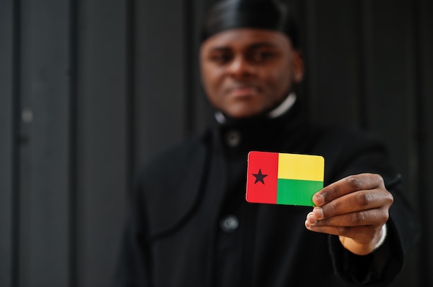 Afrikanischer Mann tragen schwarze Durag halten Guinea-Bissau-Flagge zur Hand isolierte dunkle Wand.