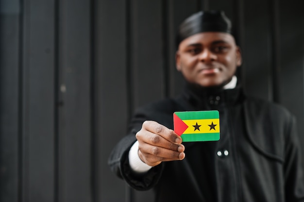 Afrikanischer Mann tragen schwarzen Durag halten Sao Tome und Principe Flagge zur Hand isolierte dunkle Wand.