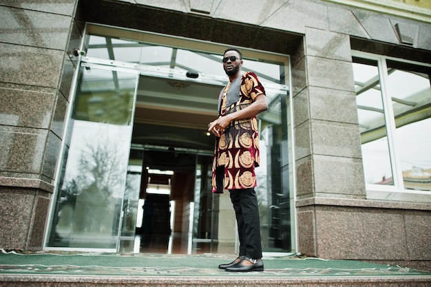 Afrikanischer stilvoller und gutaussehender Mann in traditioneller Kleidung und Sonnenbrille im Freien.
