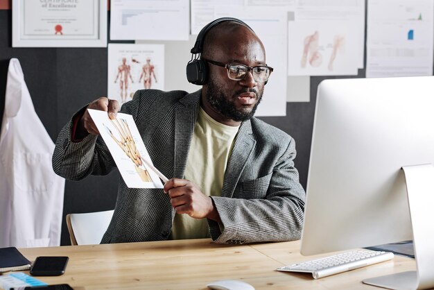 Afrikanischer Traumatologe mit Kopfhörern, der auf ein Bild mit Körperteilen zeigt und online unter Verwendung von c unterrichtet