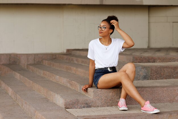 Afrikanisches Mädchen in einem weißen T-Shirt und einer Brille sitzt auf den Stufen. Sommer