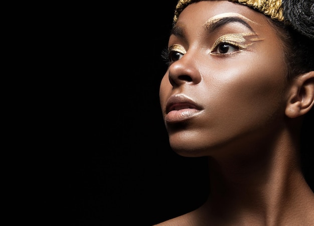 Afrikanisches Mädchen mit hellem Make-up und kreativen goldenen Accessoires auf dem Kopf Schönheitsgesichtsbild, das im Studio auf schwarzem Hintergrund aufgenommen wurde