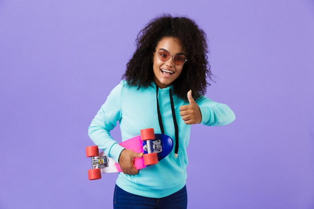 Afrikanisches Mädchen posiert isoliert über violettem Raum, der Skateboard hält.