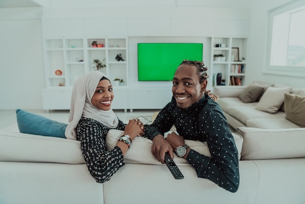 Afrikanisches Paar, das auf dem Sofa sitzt und gemeinsam fernsieht, Chroma-Green-Screen-Frau mit islamischem Hijab