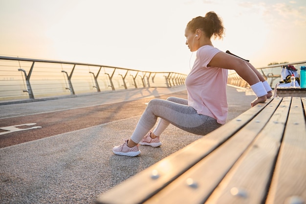 Afro-americana determinada jovem desportista fazendo exercícios no tríceps, flexionando os braços em um banco de madeira enquanto pratica um treinamento de peso corporal no ambiente urbano