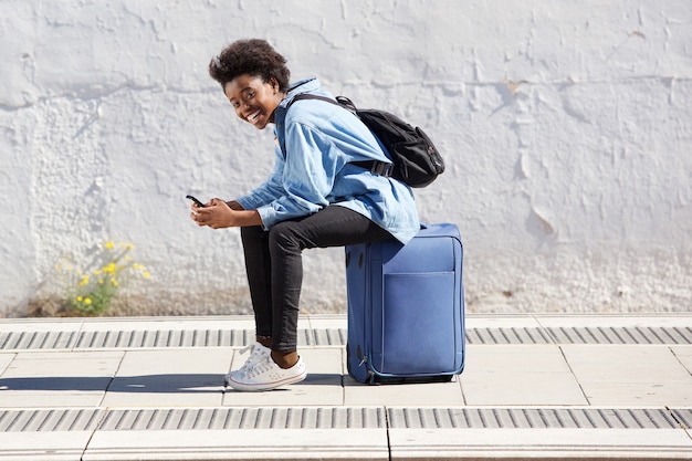 Afro americana jovem, sentando, ligado, mala, em, plataforma estrada ferro, com, telefone móvel