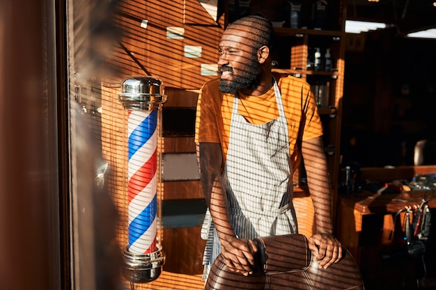 Afro-americano barbudo parado perto da janela da barbearia