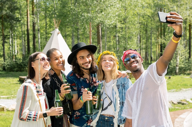 Afro-americano bonito sorridente com óculos escuros posando com os melhores amigos multiétnicos no acampamento enquanto tiravam uma selfie juntos