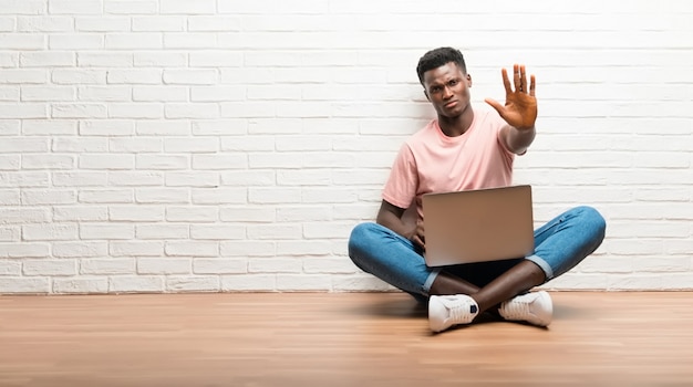 Afro americano homem sentado no chão com seu laptop fazendo parar gesto com a mão