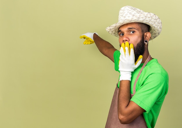 Afro-americano jovem jardineiro assustado com chapéu de jardinagem e luvas apontando para trás da boca coberta com a mão isolada na parede verde oliva com espaço de cópia