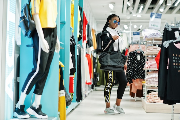 Afro-amerikanische Frauen in Trainingsanzügen und Sonnenbrillen beim Einkaufen im Sportswear-Einkaufszentrum mit Sporttasche gegen Regale Thema Sportgeschäft