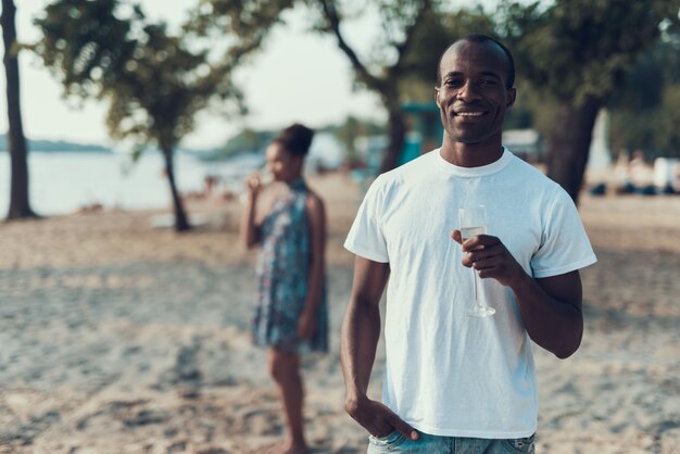 Afro casal está bebendo champanhe na praia de areia do rio.