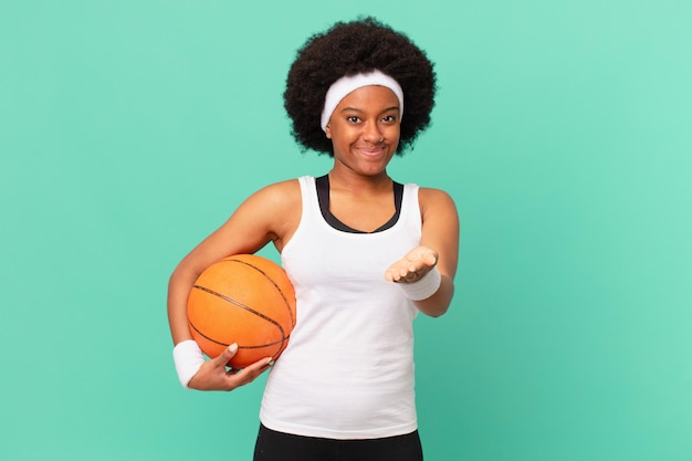 Afro-Frau, die glücklich mit freundlichem, selbstbewusstem, positivem Blick lächelt und ein Objekt oder Konzept anbietet und zeigt. Basketball-Konzept
