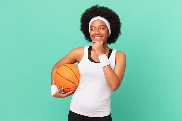 Afro-Frau, die mit einem glücklichen, selbstbewussten Ausdruck mit der Hand am Kinn lächelt, sich wundert und zur Seite schaut. Basketball-Konzept