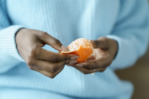 Afro Frau Hände schälen reife süße Mandarine, tragen blauen Pullover, Nahaufnahme. Winterfrucht, gesunde Ernährung, Weihnachten