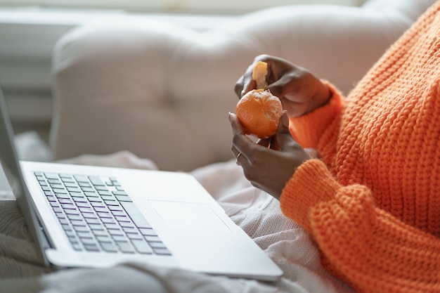 Afro Frau Hände schälen reife süße Mandarine, tragen orange Pullover, arbeiten am Laptop zu Hause